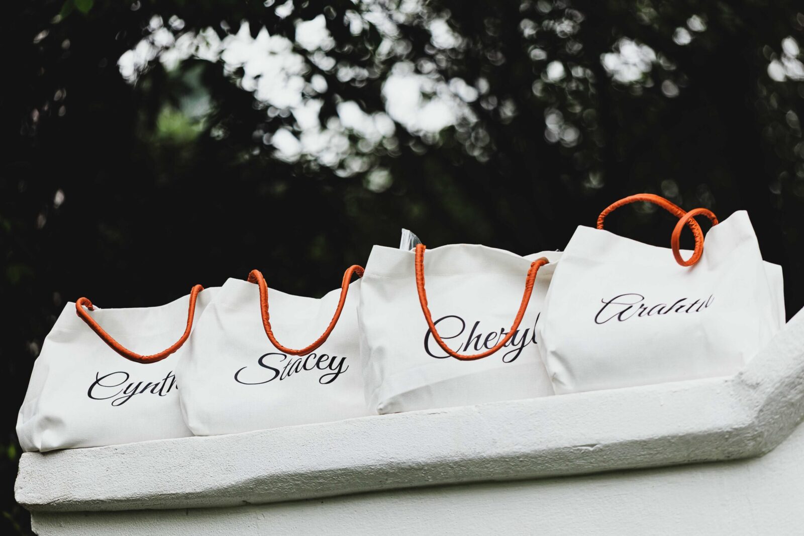 Four personalized tote bags with names and orange handles on display outdoors.